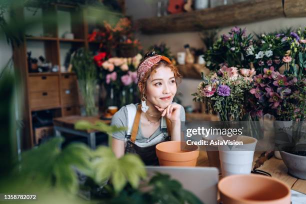 young asian female florist, owner of small business flower shop, working on laptop over counter against flowers and plants. checking stocks, taking customer orders, selling products online. daily routine of running a small business with technology - asia lady selling flower stock pictures, royalty-free photos & images