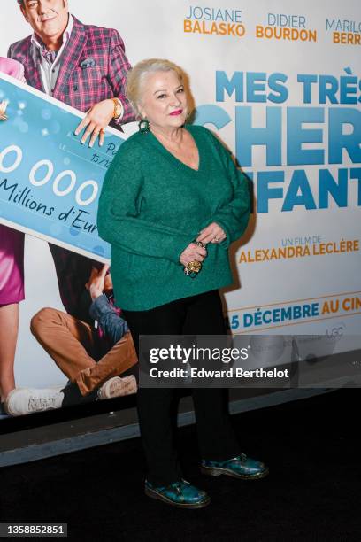 Josiane Balasko wears green bejeweled christmas earrings with pearls, a green wool v-neck pullover top, black pants, green leather shoes with printed...