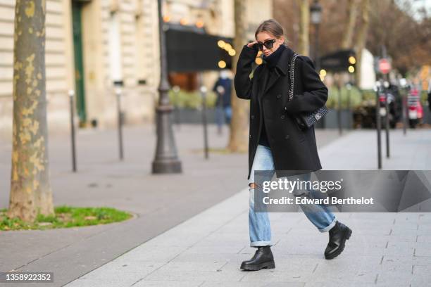 Diane Batoukina wears sunglasses from Dior, a black wool oversized turtleneck pullover, a black coat, blue faded denim ripped large jeans rolled-up...