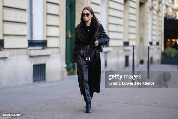 Diane Batoukina wears sunglasses, a black with white print pattern t-shirt, a black shiny leather belted long coat with a black fur collar, a black...