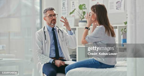 shot of a mature doctor sitting with his patient in the clinic and asking questions during a consultation - headache doctor stock pictures, royalty-free photos & images