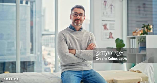 aufnahme eines reifen chiropraktikers, der allein in seiner klinik mit verschränkten armen sitzt - physiotherapist stock-fotos und bilder