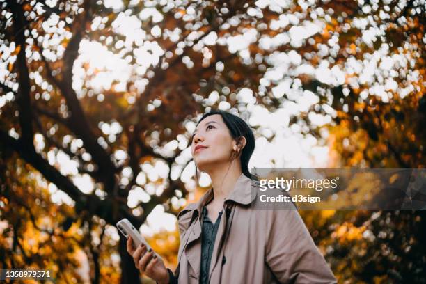 low angle portrait of young asian woman using smartphone while having a walk in the park, enjoying a beautiful autumn day outdoors and breathing fresh air. relaxing in the nature under maple trees - season in kyoto imagens e fotografias de stock