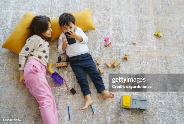 hermanos acostados en la alfombra y jugando, viendo dibujos animados en un teléfono inteligente - kids mess carpet fotografías e imágenes de stock