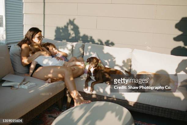 mother laying on a couch on her backyard patio with her two children and her two dogs. there is sun and shadows and everyone is relaxing and cuddling together. - mann frau hund zuhause stock-fotos und bilder