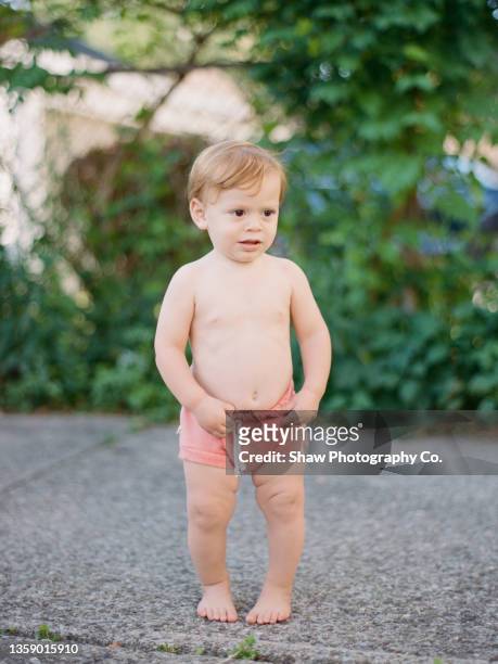 shirtless redhead toddler boy who is barefoot standing outside on the driveway looking off. his hands are touching his peach shorts and his belly button is showing. he has dark brown eyes. - belly button ストックフォトと画像