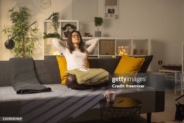mujer sentada en el sofá con las manos detrás de la cabeza después de administrar la temperatura del aire acondicionado - emoción positiva fotografías e imágenes de stock