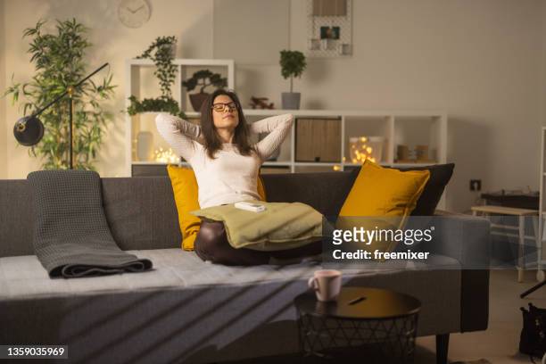 donna seduta sul divano con le mani dietro la testa dopo aver gestito la temperatura del condizionatore d'aria - condizionatore casa foto e immagini stock