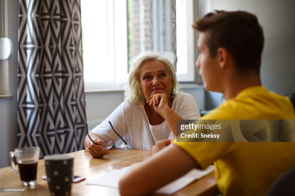 A teenager is sitting with his granny at the table, talking. Grandson is doing his homework and his granny is helping him.