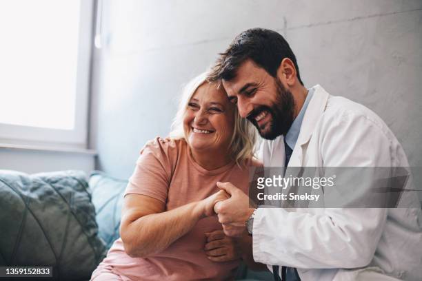 若い医者と先輩の女性がソファに座っています。二人とも笑顔です。医者が先輩女性の手を握っている。 - patient ストックフォトと画像