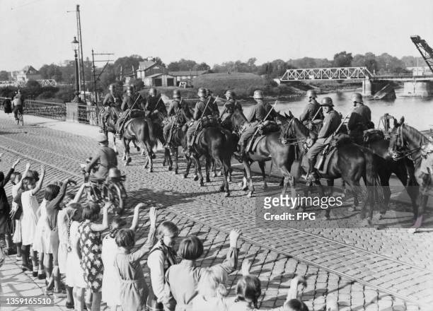 Group of young children wave as a German Wehrmacht cavalry column from the 1st Cavalry Brigade on the march during military manoeuvres passes over a...
