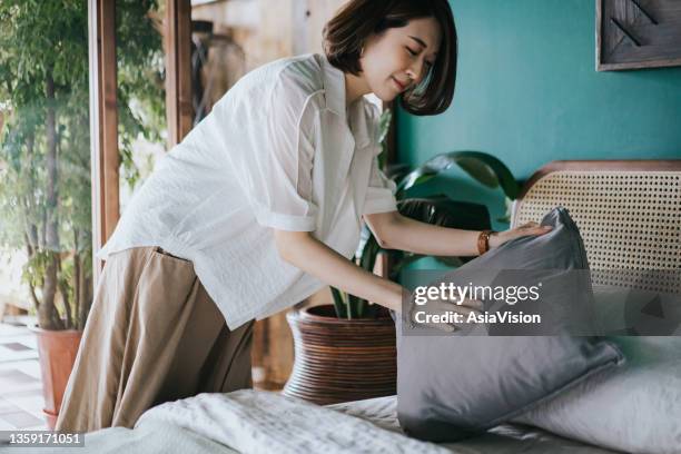 young asian woman doing her morning routine, arranging pillows and making up her bed at home. let's get the day started - making bed stockfoto's en -beelden