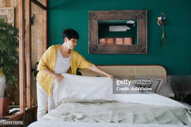 senior asian woman doing her morning routine, making up her bed at home. let's get the day started - bed linen stockfoto's en -beelden