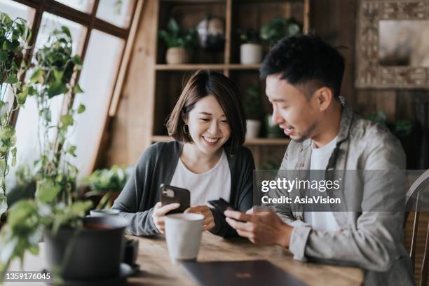 happy young asian couple having a coffee date in cafe. drinking coffee and chatting while using smartphone together. enjoying a relaxing moment - happy couple using cellphone stockfoto's en -beelden