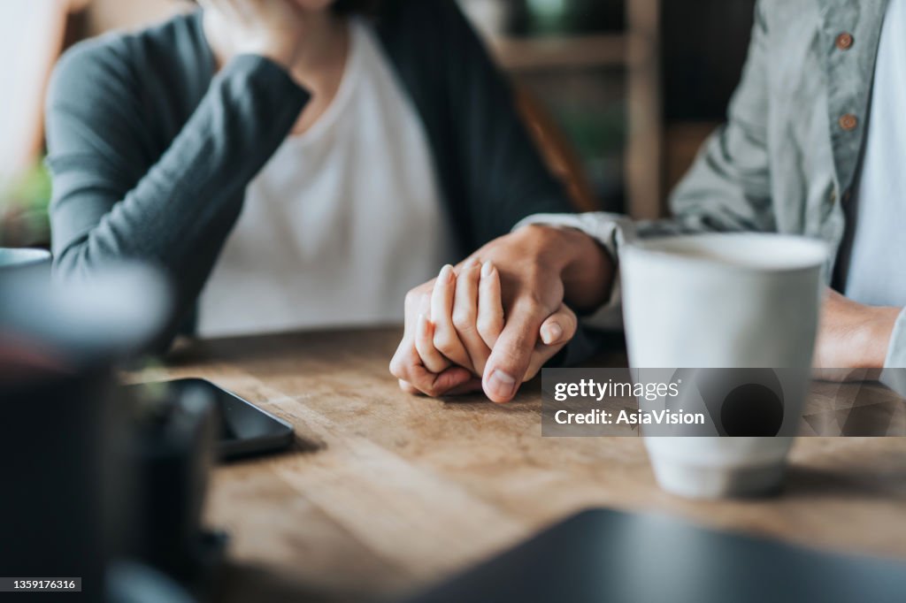 Close up of young Asian couple on a date in cafe, holding hands on coffee table. Two cups of coffee and smartphone on wooden table. Love and care concept