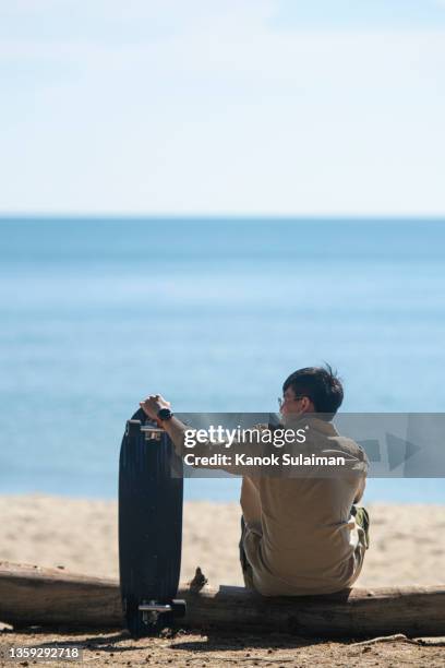 asian man with surfskate resting on the beach - asian man seated stockfoto's en -beelden