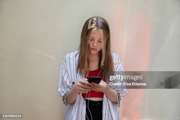 teenage girl using mobile phone and leaning against a white painted wall, with red reflections bounced off from the (out-of-frame) rear lights of a nearby vehicle, projected onto the wall - one teenage girl only stockfoto's en -beelden