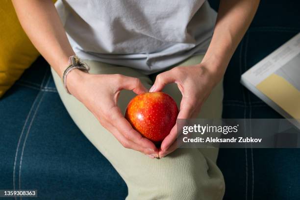 a red apple in the hands of a woman who is sitting at home on a bright sofa. a girl eats a fruit. cultivation of organic agricultural products. the concept of vegetarian, vegan, raw food and diet. proper healthy nutrition. hands in the shape of a heart. - healthcare worker beauty in nature stock pictures, royalty-free photos & images