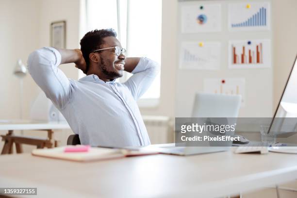 afro-american businessman holding hands behind head while sitting at office - relief emotion stock pictures, royalty-free photos & images