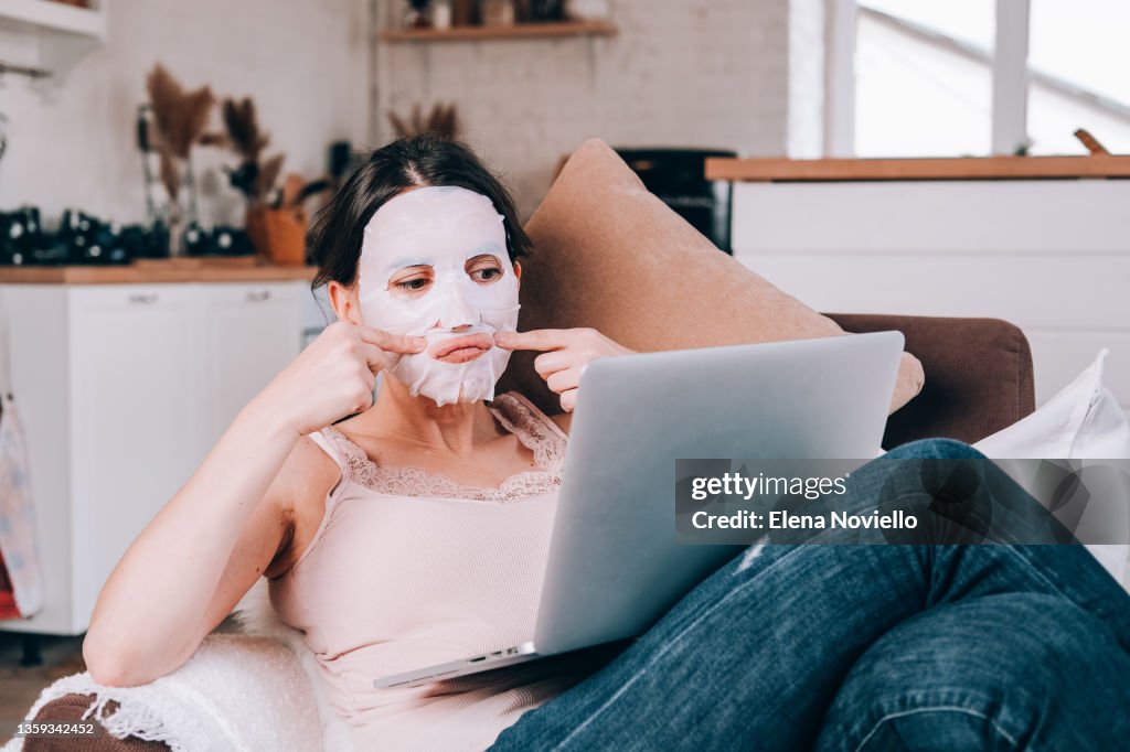 Facial massage. woman in a moisturizing mask massaging her face. A woman sits in a cozy chair and watches online course on facial skin care. face fitness