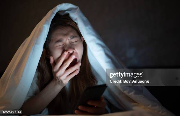woman yawning while playing and surfing internet on her smartphone late at night on bed. - midnight stock pictures, royalty-free photos & images