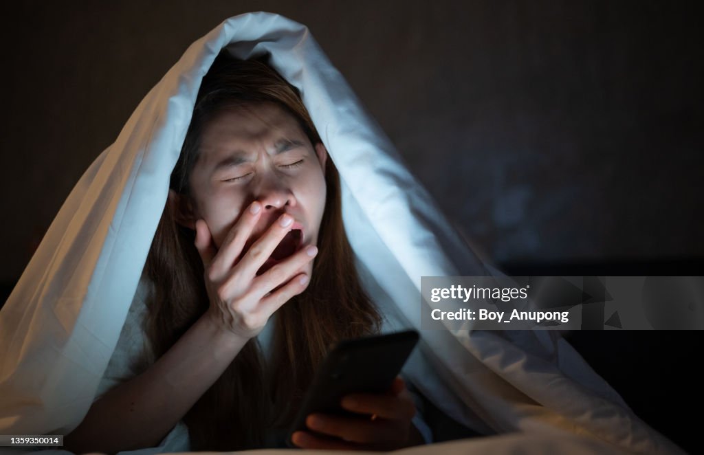 Woman yawning while playing and surfing internet on her smartphone late at night on bed.