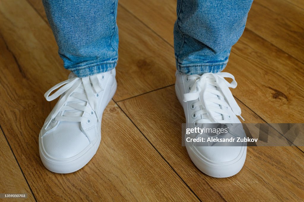 The girl's legs are in new clean white modern sneakers and jeans. A woman or a teenager in sports shoes stands on the wooden floor of a house or indoors. A fashionable and stylish lifestyle. A copy of the text space.