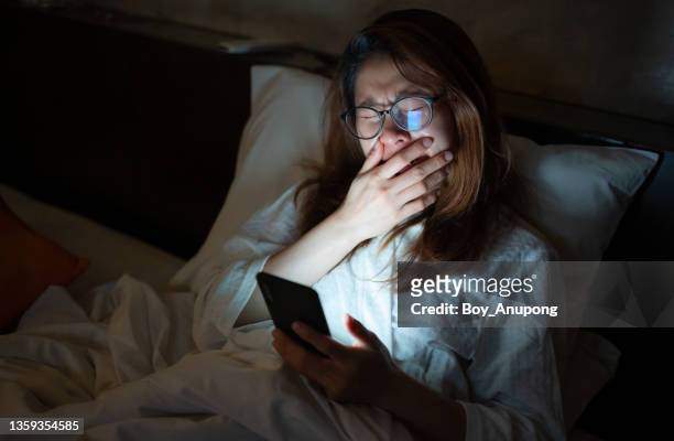 woman yawning while playing and surfing internet on her smartphone late at night on bed. - yawning stock photos et images de collection