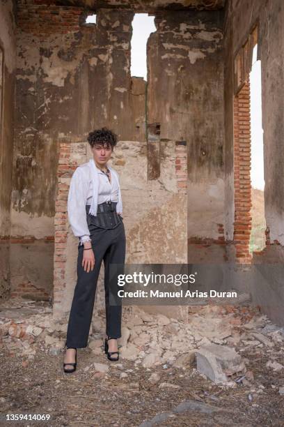 non-binary guy with curly hair, vintage clothes and heels looking at the camera in an abandoned factory. - androgyn stock pictures, royalty-free photos & images
