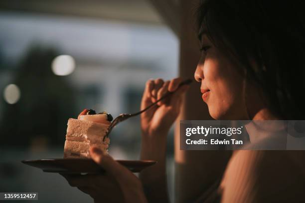asian chinese beautiful woman enjoying slice of cake at home during sunset looking away through window curtain - evening indulgence stock pictures, royalty-free photos & images