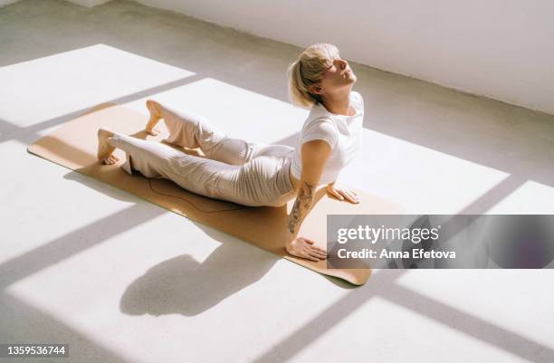 beautiful authentic woman with tattoos and short blond hair is doing yoga in upward facing dog position on yoga mat in front of a window. she is wearing a light-colored casual clothing. concept of relaxation exercises - practicing stock-fotos und bilder