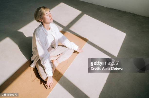 beautiful authentic woman with short blond hair is meditating sitting in lotus position on yoga mat in front of a window. she is wearing a light-colored casual clothing. concept of relaxation exercises - blonde yoga foto e immagini stock