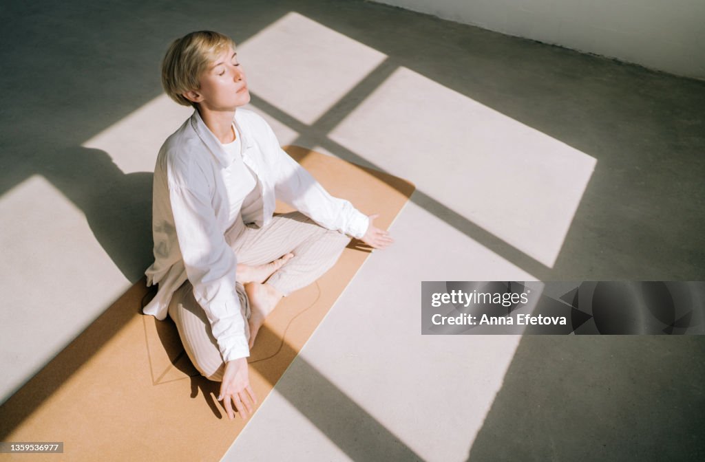 Beautiful authentic woman with short blond hair is meditating sitting in lotus position on yoga mat in front of a window. She is wearing a light-colored casual clothing. Concept of relaxation exercises