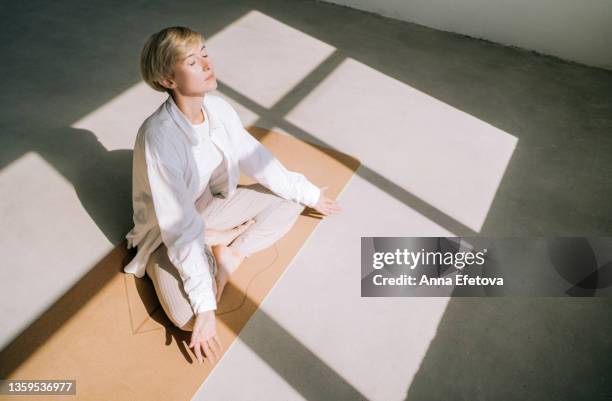 beautiful authentic woman with short blond hair is meditating sitting in lotus position on yoga mat in front of a window. she is wearing a light-colored casual clothing. concept of relaxation exercises - ioga imagens e fotografias de stock