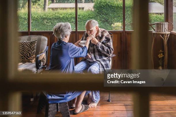 female caretaker feeds feeble elderly man on screened porch - parkinsons disease stock pictures, royalty-free photos & images
