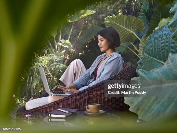 asian woman working from home sitting in garden furniture surrounded by tropical plants, using laptop computer - tropical elegance stock pictures, royalty-free photos & images