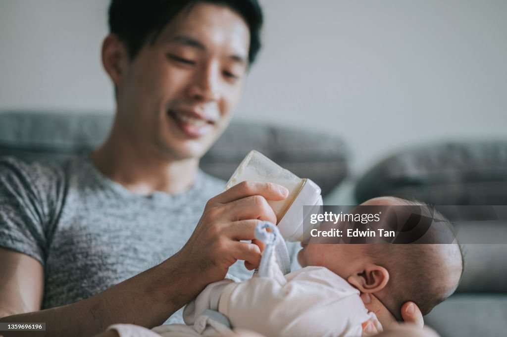 Asiatisch-chinesischer junger Vater füttert seinen kleinen Sohn am Wochenende im Wohnzimmer mit Milchflasche
