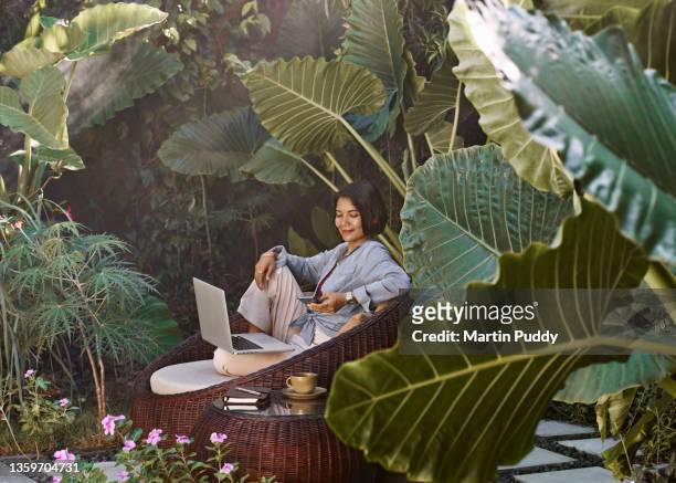 asian woman working from home sitting in garden furniture surrounded by tropical plants, using laptop computer and mobile phone - low key stock-fotos und bilder