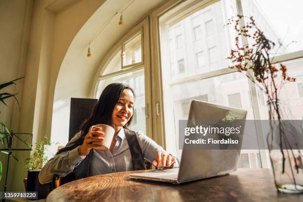 a middle-aged asian woman in blue jeans sitting on the bed in a yoga pose in front of a laptop - banking document stock-fotos und bilder