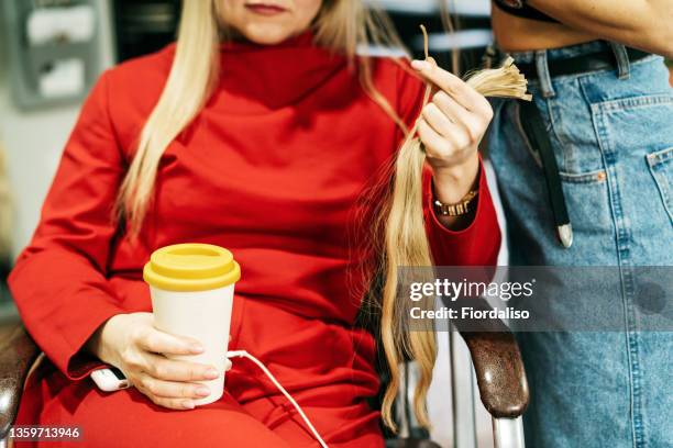 middle-aged blonde in a red suit sitting in a chair with a bamboo reusable coffee cup in her hands, handing a strand of hair to the stylist for extensions - alongamento de cabelo - fotografias e filmes do acervo