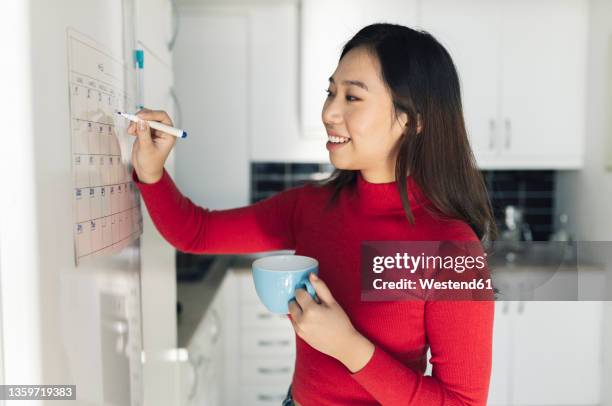 smiling woman with coffee cup writing on calendar in kitchen - calendario español fotografías e imágenes de stock