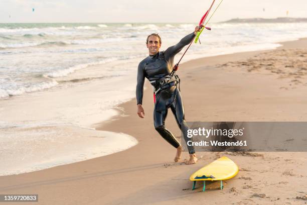 smiling man holding rope of parachute by kiteboard at beach - kiteboarding fotografías e imágenes de stock