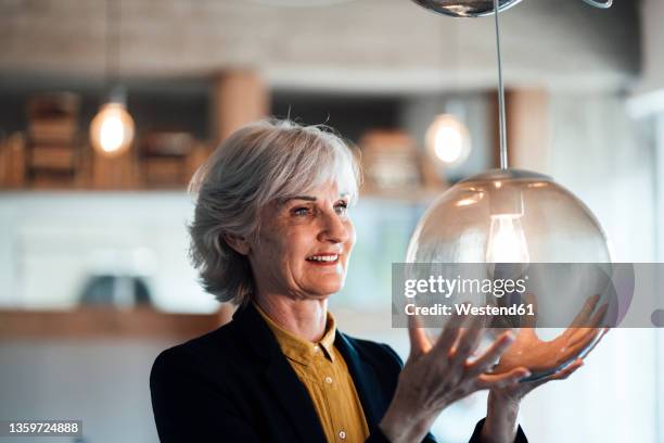 smiling businesswoman looking at illuminated pendant light in office - lightbulb stock-fotos und bilder