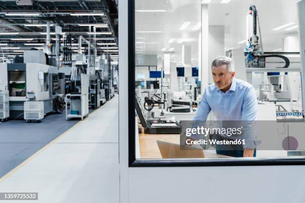 confident businessman looking through glass at factory - lean manufacturing stockfoto's en -beelden