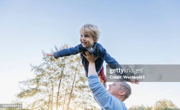 smiling man holding aloft blond boy flying under clear sky - 飛行機のまね ストックフォトと画像