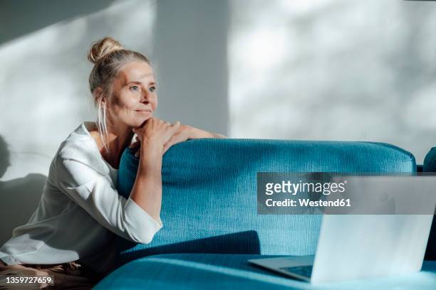 thoughtful woman with laptop leaning on blue sofa at home - casual woman pensive side view stockfoto's en -beelden