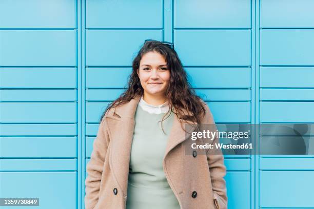 smiling curvy woman wearing overcoat in front of blue wall - kräftig gebaut stock-fotos und bilder