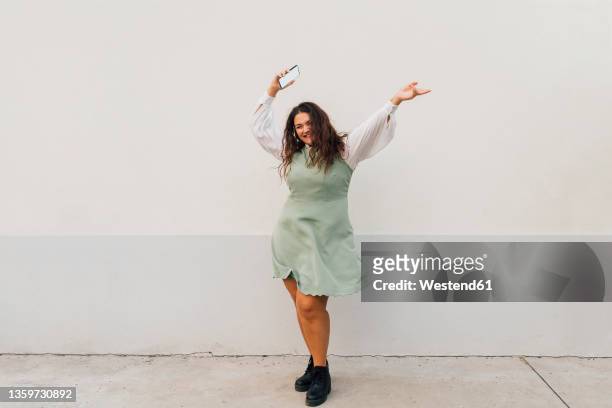 cheerful plus size woman with smart phone dancing against wall - fat woman dancing stockfoto's en -beelden