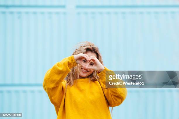 smiling woman looking through heart sign in front of blue wall - gestikulieren mit den händen stock-fotos und bilder