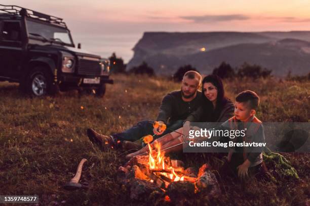 family enjoying outdoors on picnic near fire in evening. and baking corn - family camping stock-fotos und bilder